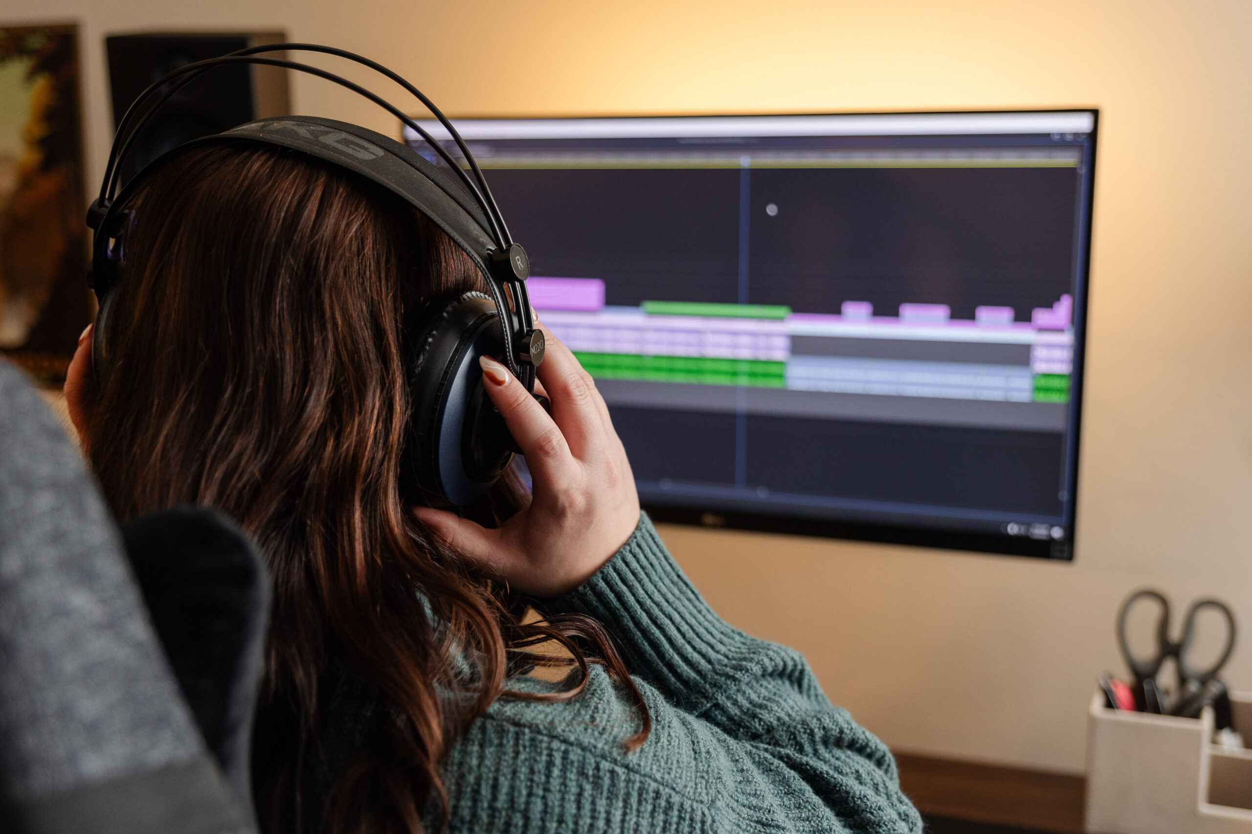 a woman with brown hair sits in front of her video editing console, putting on her headphones ready to get to work
