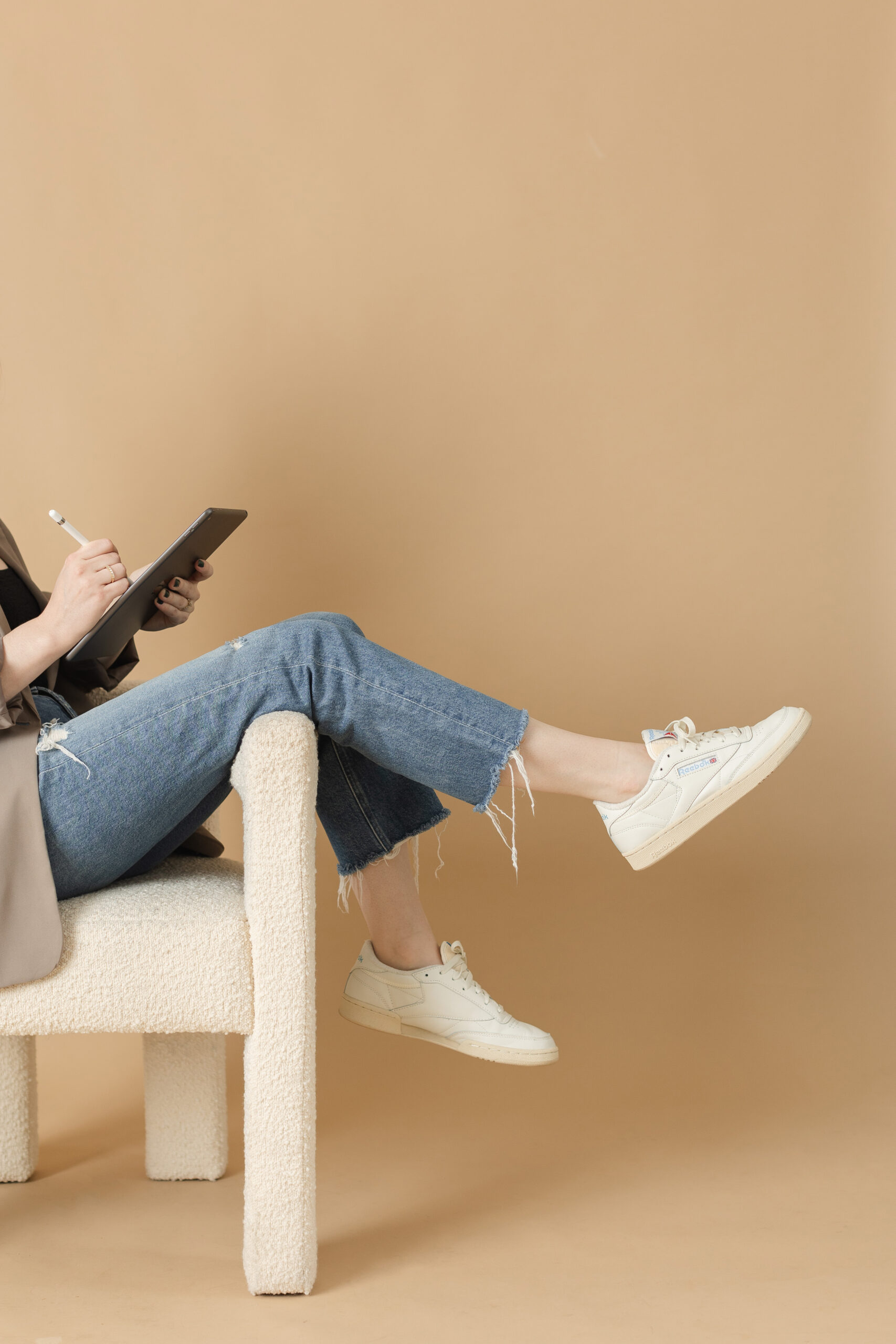 woman web designer sits, drawing on her ipad, with legs draped over the arm of a cool chair in ripped jeans and white sneakers. she seems really cool