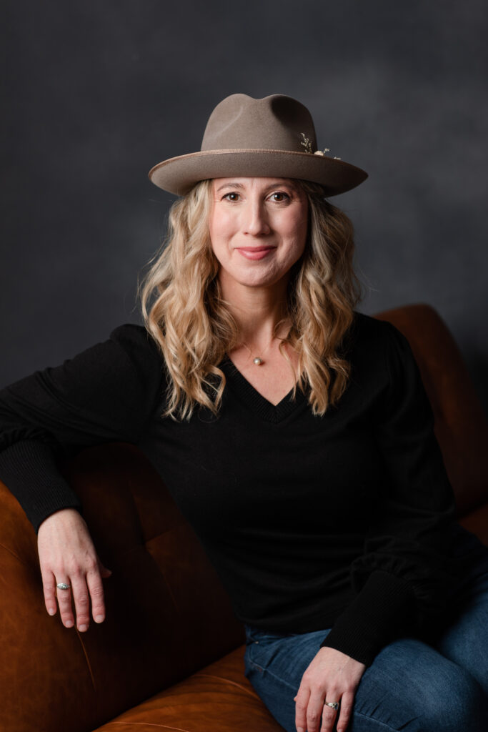 headshot: a blonde haired woman wearing a cool hat and a black blouse sits casually on a leather couch