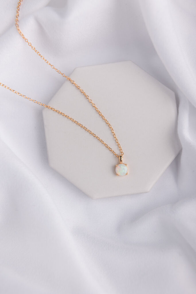 a necklace with an opal stone on a gold chain rests on a white stone tile, surrounded by lush white fabric