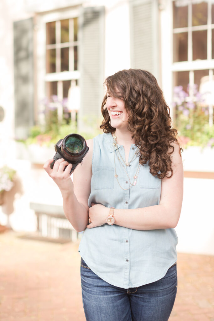 a curly haired woman in a chambray blue shirt smiles to someone off camera while holding a professional photo camera