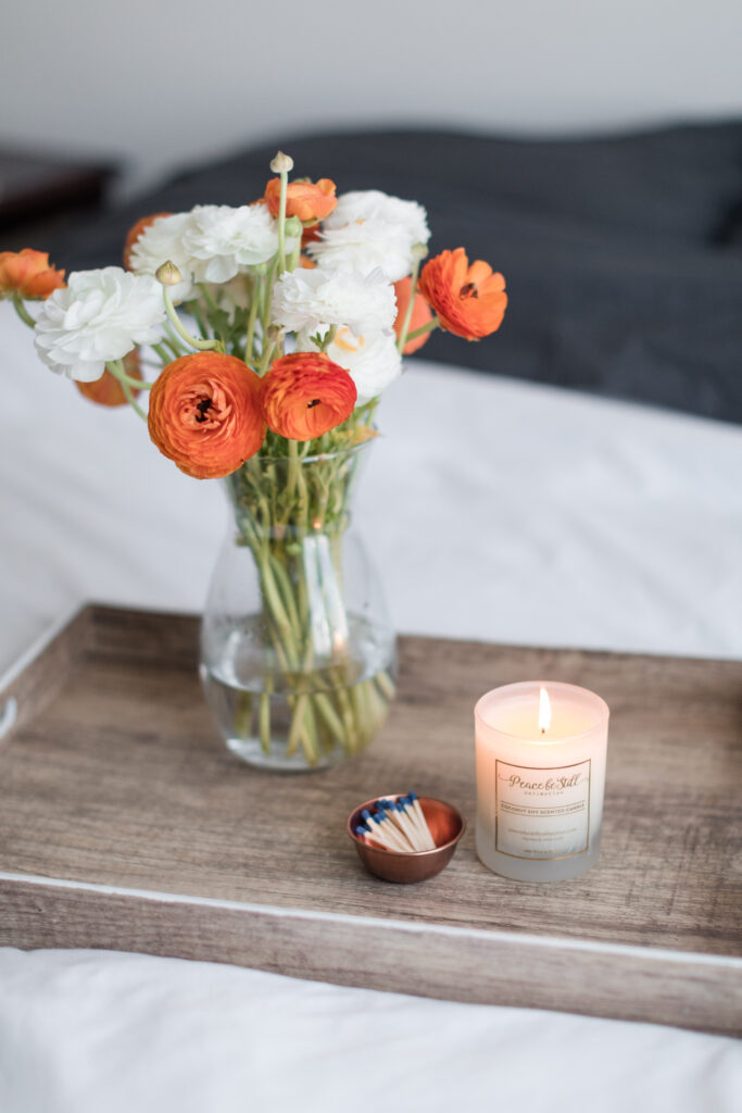 a candle rests on a tray along with orange and white flowers on a bed draped in a cozy white comforter