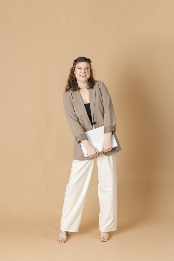 a brown haired woman in a tan blazer and white slacks holds her laptop playfully in her hands in front of a plain tan background