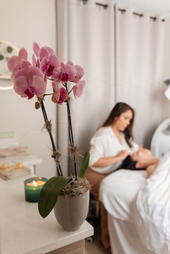 a purple orchid fills the frame, while in the background we see a woman receiving a facial massage