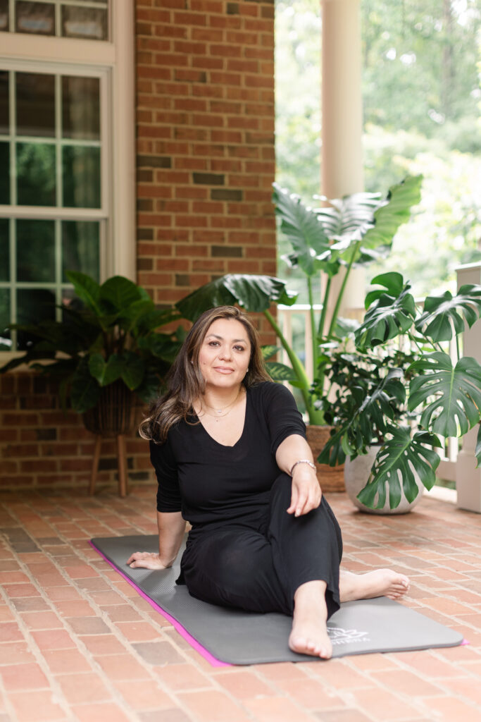 a spa owner wearing all black sits casually on a yoga may, surrounded by lush green plants