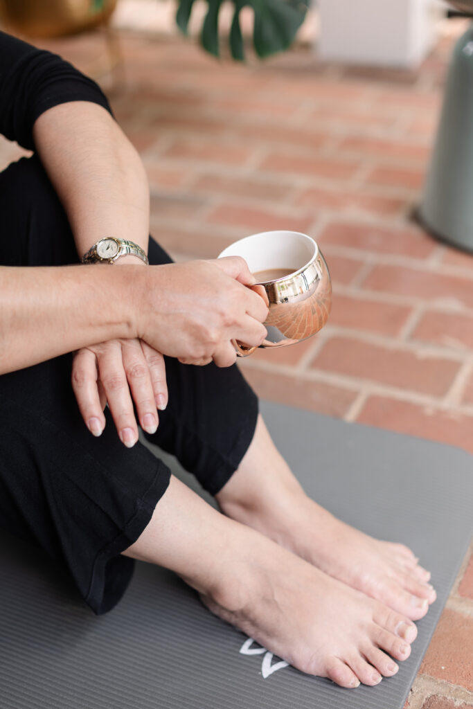 a close up shot of a woman's hands and feet while seated on a yoga mat sipping hot tea