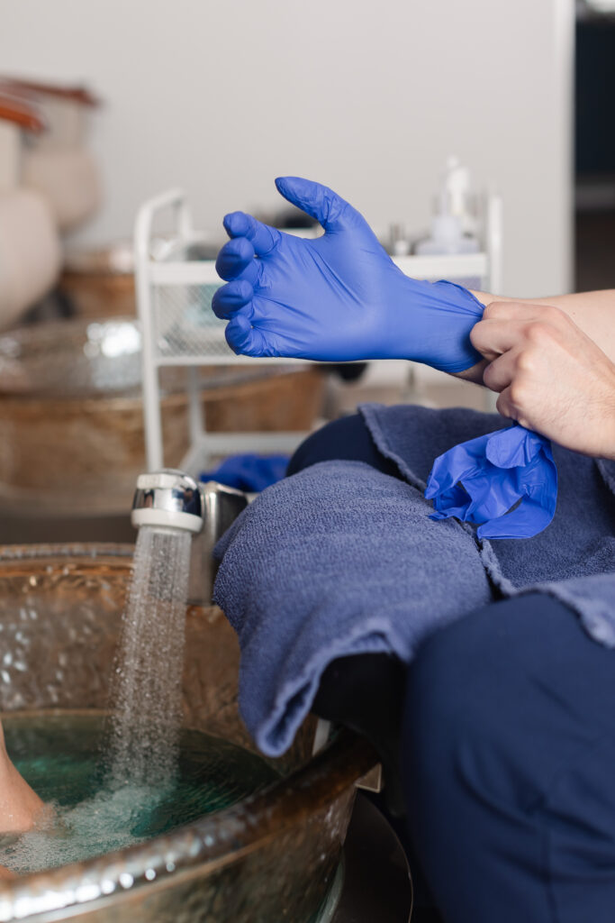 a nail technician puts on his blue gloves as he fills the pedicure bowl with water.