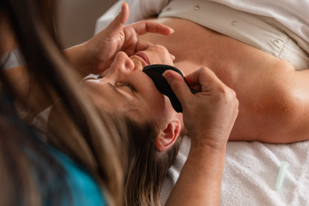 a woman lays on a massage bed, receiving a facial gua sha massage at a luxury spa