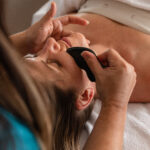 a woman lays on a massage bed, receiving a facial gua sha massage at a luxury spa