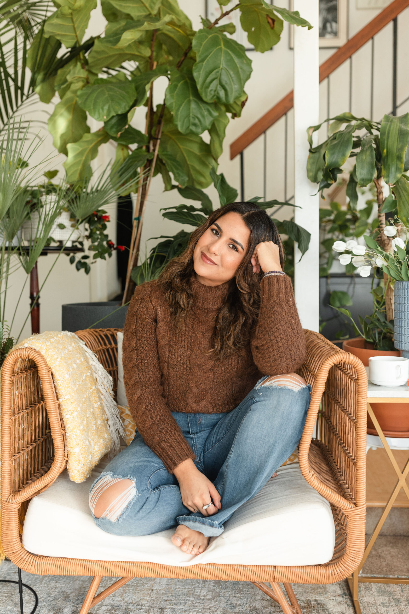 a latina photograper sits curled up on a rattan chair, surrounded by plants