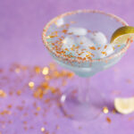 an ice cold margarita glass surrounded by sparking gold confetti sits in front of a purple backdrop