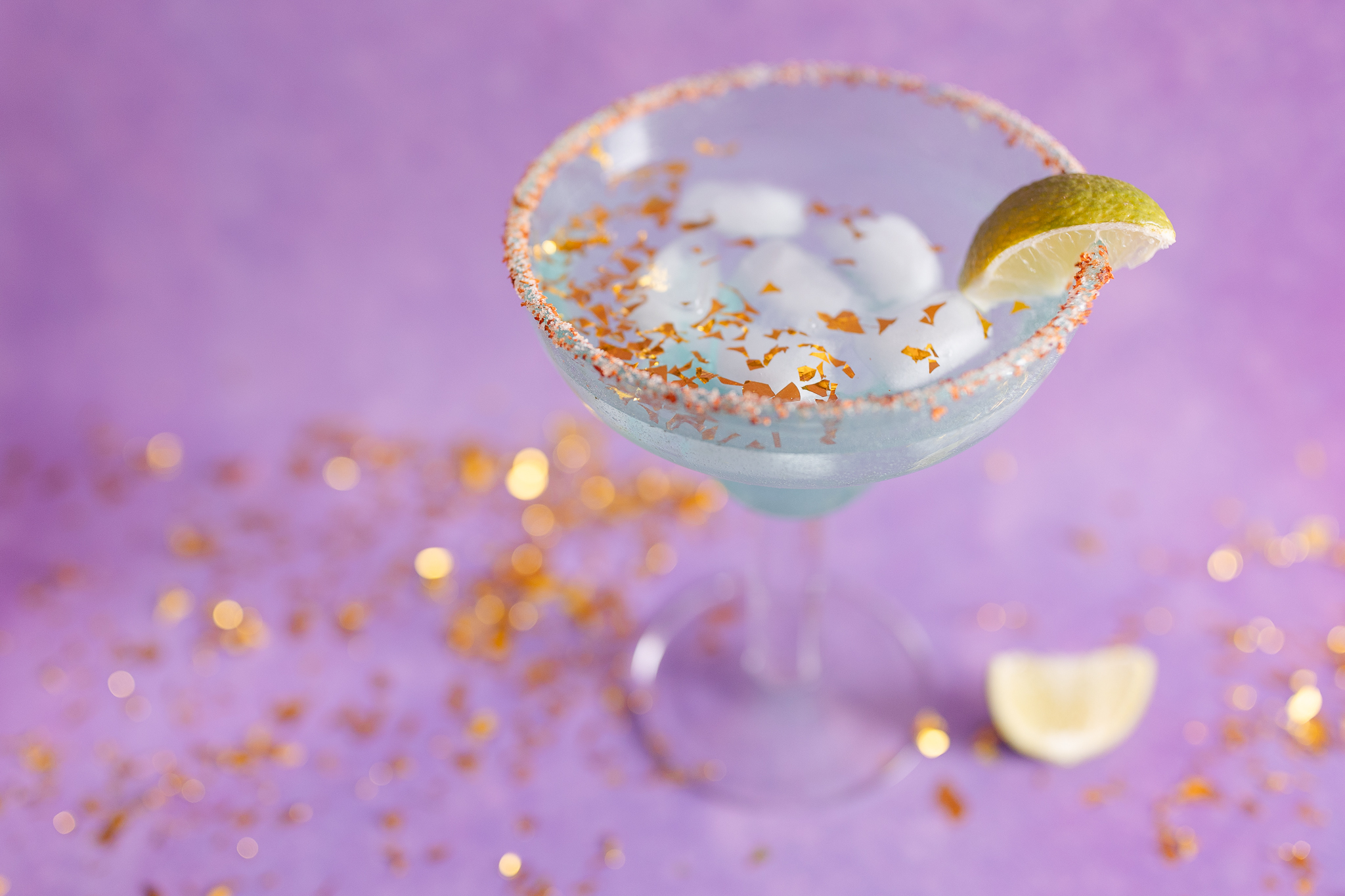 an ice cold margarita glass surrounded by sparking gold confetti sits in front of a purple backdrop