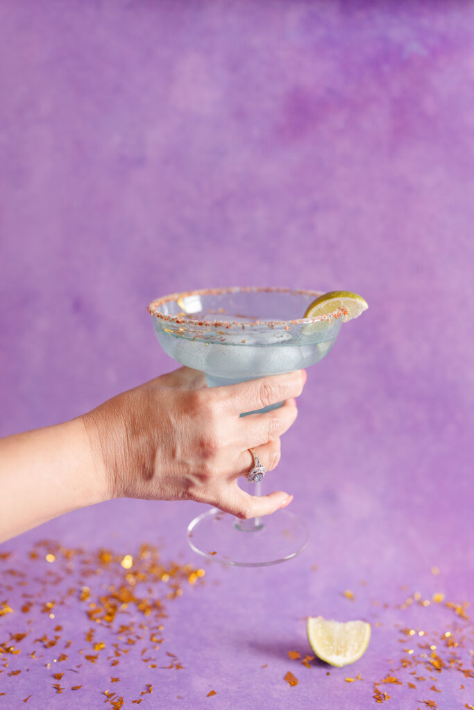 a woman holds a margarita glass in front of a colorful purple backdrop, surrounded by gold confetti