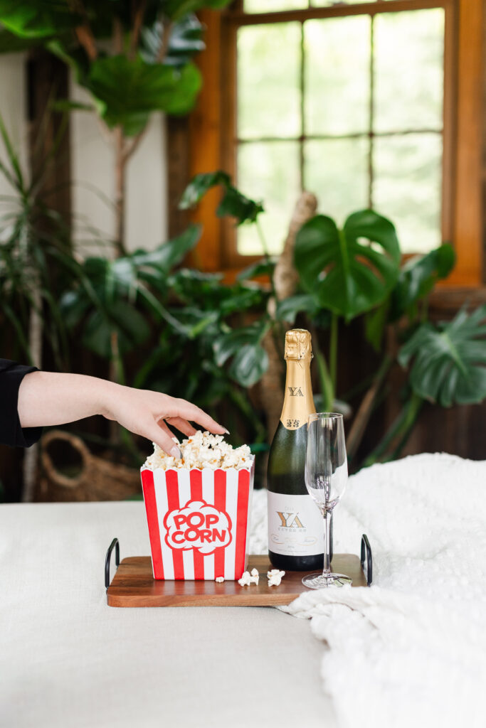 a woman reaches into a tub of popcorn with a glass of champagne nearby