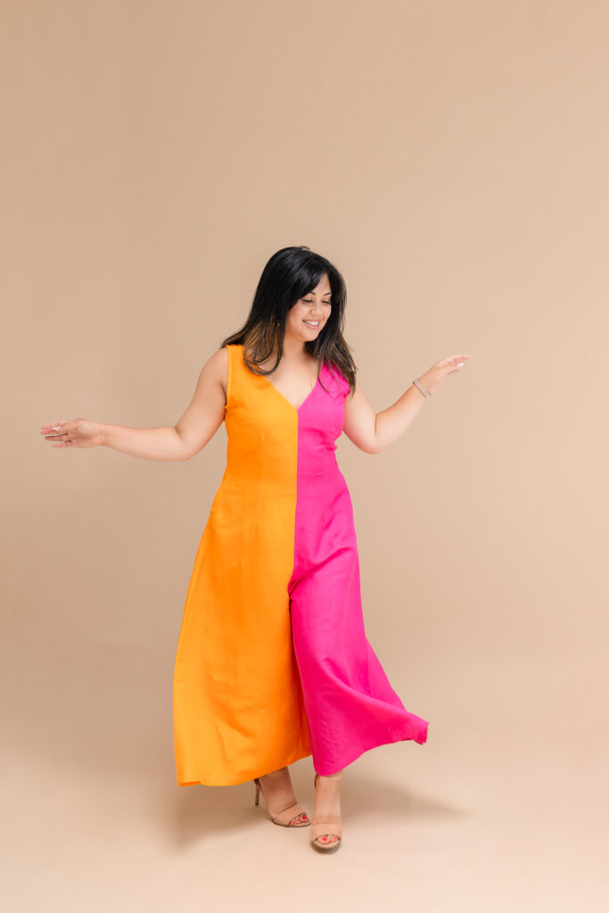 a grinning woman twirls on a beige backdrop in a magenta and orange two-tone jumpsuit