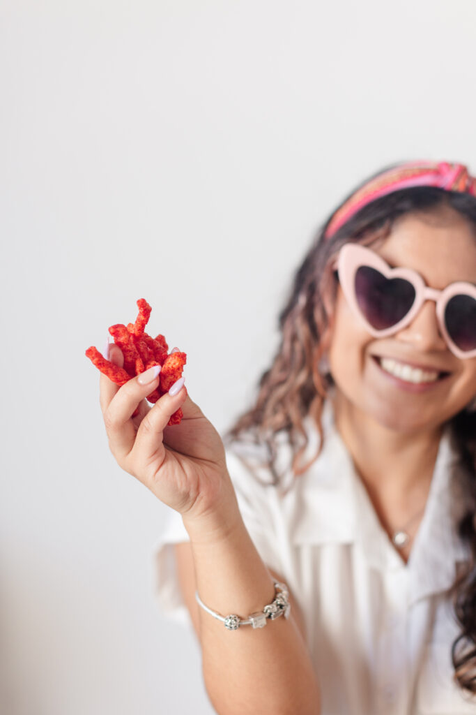 a smiling woman with pink heart shaped sunglasses holds a handful of hot cheetos