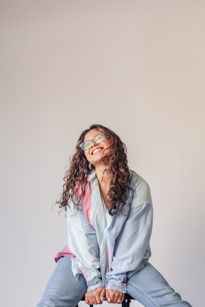 a grinning curly-haired young woman sits on a stool in a colorful shirt